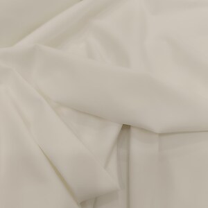 Op de afbeelding: Close-up van een witte stof met zachte plooien en subtiele schaduwen. De stof lijkt glad en mogelijk licht van gewicht, wat suggereert dat deze gebruikt kan worden voor kleding of interieurdecoratie.