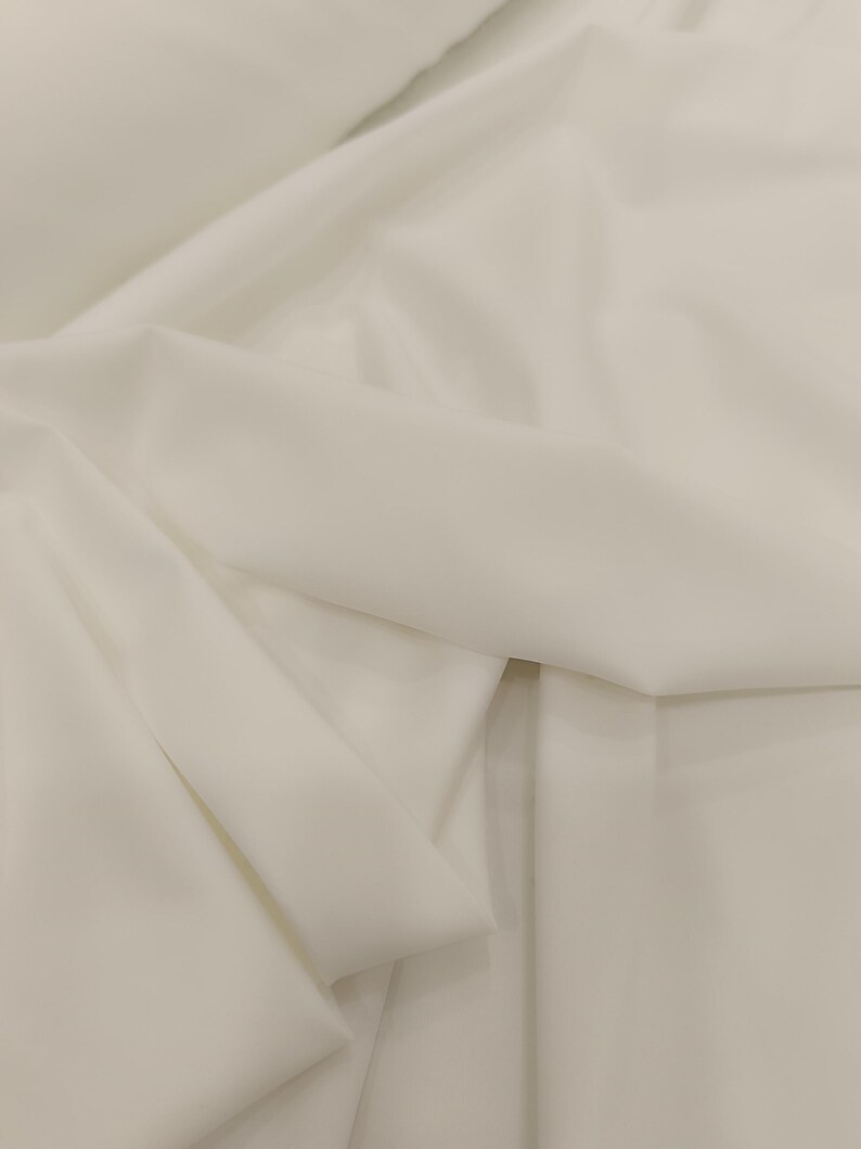 Op de afbeelding: Close-up van een witte stof met zachte plooien en subtiele schaduwen. De stof lijkt glad en mogelijk licht van gewicht, wat suggereert dat deze gebruikt kan worden voor kleding of interieurdecoratie.