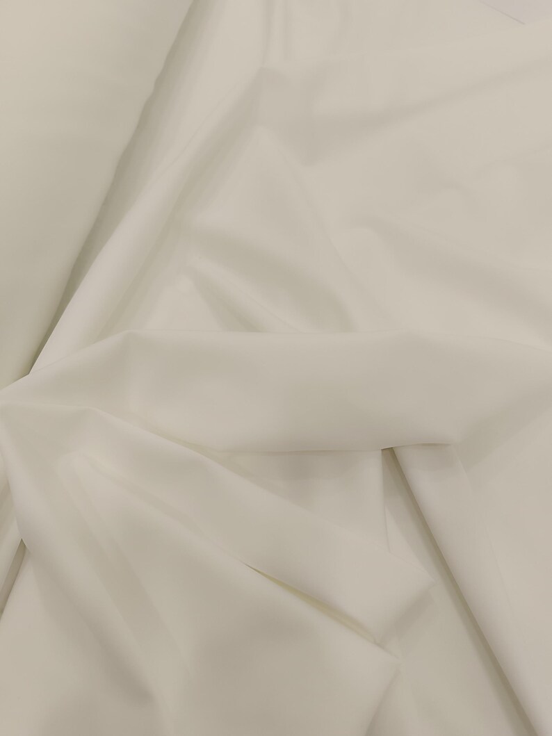 Op de afbeelding: Close-up van een witte stof met zachte plooien en subtiele schaduwen. De stof lijkt glad en licht, mogelijk geschikt voor kleding of gordijnen. De algehele toon is neutraal en schoon.