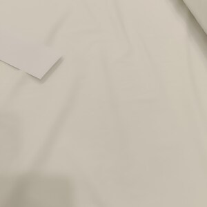 Op de afbeelding: Een close-up van een rol off-white stof. De stof is glad en lijkt een effen kleur te hebben. Een klein, gevouwen stuk van dezelfde stof bevindt zich in de linkerbenedenhoek.