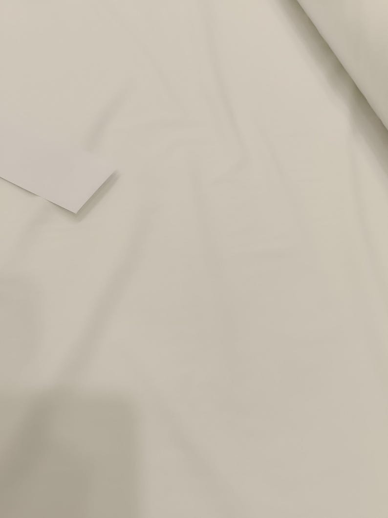 Op de afbeelding: Een close-up van een rol off-white stof. De stof is glad en lijkt een effen kleur te hebben. Een klein, gevouwen stuk van dezelfde stof bevindt zich in de linkerbenedenhoek.