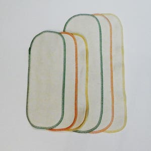 Peut inclure: Quatre lingettes pour bébé en tissu blanc avec des coutures vertes, orange et jaunes autour des bords.