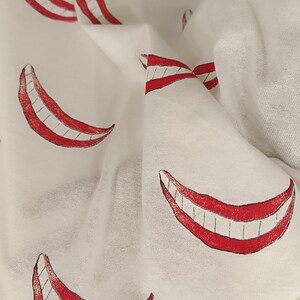 Może przedstawiać: Biała tkanina z powtarzającym się wzorem czerwonych uśmiech&oacute;w, każdy z czarnymi konturami i białymi zębami. Materiał jest miękki i lekki, z widocznymi fałdami i zagnieceniami, sugerującymi szalik lub podobny przedmiot.