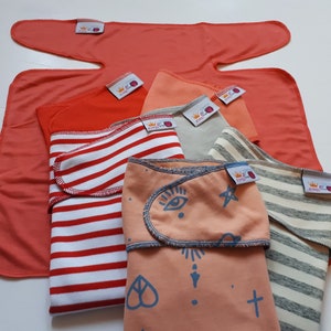 Può includere: Un assortimento di fasce per neonati in vari colori e fantasie. Le fasce includono corallo tinta unita, strisce rosse e bianche e pesca con illustrazioni blu. Ogni fascia ha un'etichetta con un logo a corona.