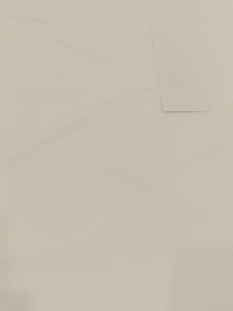 Op de afbeelding: Een effen, gebroken witte achtergrond met subtiele kleurschakeringen. Een rechthoekig stuk lichtgekleurd papier is gedeeltelijk zichtbaar in de rechterbovenhoek, waardoor een zachte schaduw ontstaat. De compositie is eenvoudig en minimalistisch.