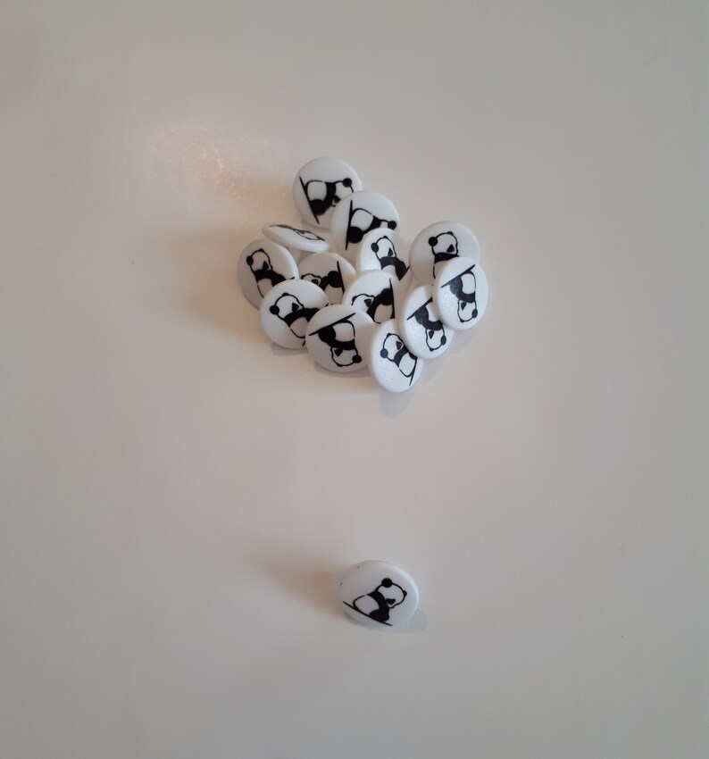 May include: A group of white round buttons with black panda bear designs, scattered on a white surface. One button is separated from the group.