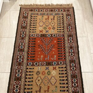 May include: A traditional rug with a brown, red, and blue geometric pattern. The rug has a fringe border and is made of wool.