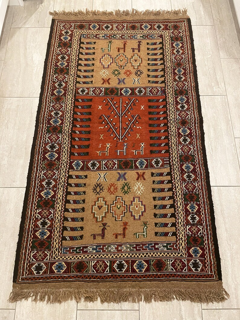 May include: A traditional rug with a brown, red, and blue geometric pattern. The rug has a fringe border and is made of wool.