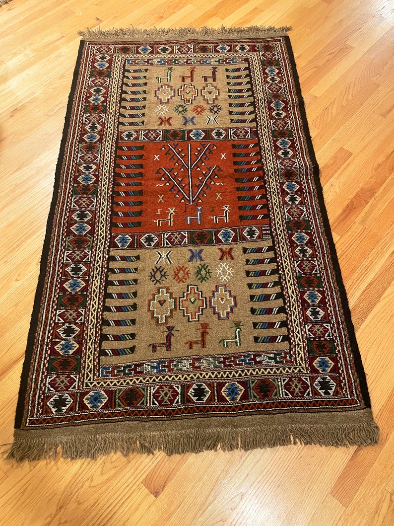 May include: A traditional rug with a brown background and intricate geometric patterns in red, blue, green, and black. The rug features a central design of a tree with stylized branches and leaves.