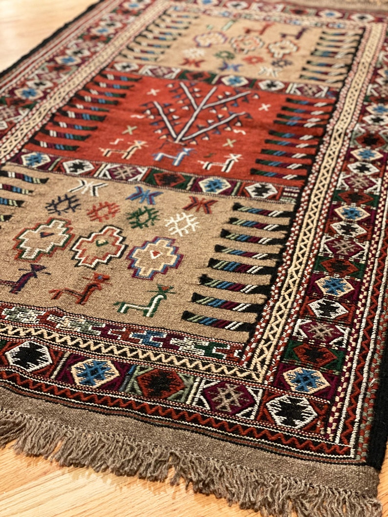 May include: A traditional rug with a beige background and intricate patterns in shades of red, blue, green, and black. The rug has a fringe border and features geometric and floral motifs.