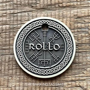 Könnte beinhalten: Ein runder, silberfarbener Metallanhänger mit dem Namen "ROLLOE" in der Mitte, umgeben von komplizierten Symbolen und einem keltischen Knotenmuster. Der Anhänger hat oben ein kleines Loch zur Befestigung. "The Barking Tag Smith" ist unten aufgedruckt.