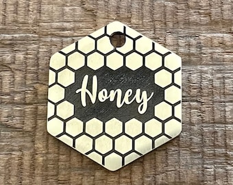 Puede incluir: Una etiqueta hexagonal dorada para mascotas con un patrón de panal y el nombre "Honey" grabado en negro.