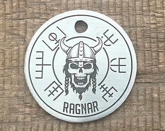 Puede incluir: Una etiqueta redonda de metal plateado con un diseño de calavera vikinga y la palabra "RAGNAR" grabada en la parte inferior. La calavera lleva un casco con cuernos y tiene el pelo trenzado. La etiqueta tiene un agujero en la parte superior para sujetar una cadena o una cuerda.