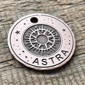 Puede incluir: Una etiqueta redonda de metal de color cobre con un diseño de brújula y la palabra "ASTRA" grabada alrededor del borde. La etiqueta tiene un pequeño agujero en la parte superior para sujetarla a un llavero u otro objeto.