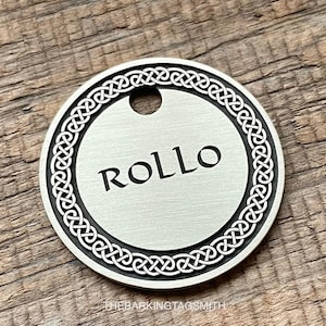 May include: A round, silver-colored pet tag with the name "ROLLO" in black, centered on the tag. The tag has a decorative Celtic knot border and a hole at the top for attachment. The tag is made of metal.