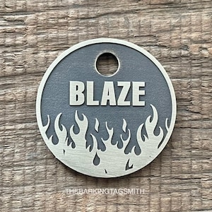 Puede incluir: Una chapa redonda para mascotas de color plateado con el nombre "BLAZE" en letras blancas en negrita. La chapa presenta un diseño de llamas alrededor del borde y un agujero en la parte superior para sujetarla. El texto "THEBARKINGTAGSMITH" está en la parte inferior.