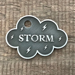 May include: A silver metal cloud-shaped tag with the word "STORM" in white lettering and lightning bolt designs around the edge.