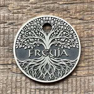 May include: A silver-colored, circular pendant featuring a detailed tree design with the word "FREYJA" in the center. The pendant has a hole at the top and intricate root details at the bottom. The Barking Tag Smith is printed at the bottom.