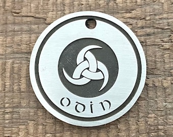 Puede incluir: Un colgante redondo de plata con un símbolo triquetra negro y la palabra "odin" inscrita debajo.