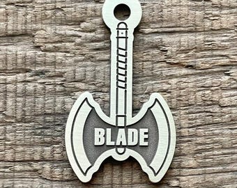May include: A silver axe-shaped pendant with the word "BLADE" in the centre. The pendant has a loop at the top for attachment and is set against a wooden background. The text "THEBARKINGTAGSMITH" is at the bottom.