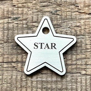 May include: A silver star-shaped dog tag with the word "STAR" engraved on it. The tag has a hole at the top for attaching to a collar. The tag is on a wooden surface.