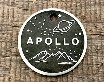 May include: A black and silver circular tag with the word "APOLLO" in white, along with a mountain range, stars, and a planet design. The tag has a small hole at the top and is set against a wooden background.