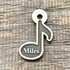 Könnte beinhalten: Ein silberner Musiknoten-Hundeanhänger aus Metall mit dem eingravierten Namen "Miles". Der Anhänger hat oben ein Loch zum Befestigen an einem Halsband.