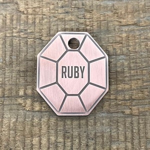 Può includere: Un'etichetta per cani ottagonale di colore rame con un bordo smussato e la parola "RUBY" incisa al centro.