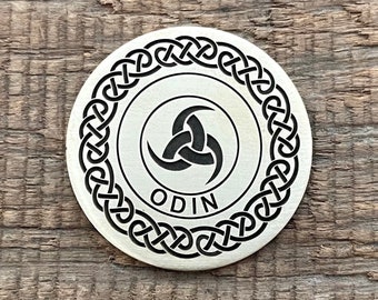 Puede incluir: Una moneda redonda de metal de color latón con un borde negro de nudo celta y un símbolo triquetra negro en el centro. La palabra "ODIN" está inscrita debajo del símbolo.