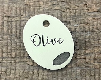 Puede incluir: Una etiqueta de identificación ovalada blanca con el nombre "Olive" grabado en letras negras. La etiqueta tiene un pequeño agujero en la parte superior para sujetarla a un collar.