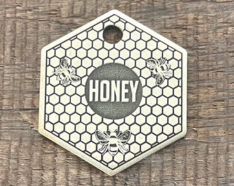 Puede incluir: Una etiqueta hexagonal de latón para mascotas con un patrón de panal y tres abejas. La etiqueta tiene un círculo redondo en el centro con la palabra "HONEY" grabada en negro.