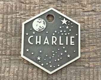 Puede incluir: Una chapa hexagonal de perro de plata con un diseño de luna y estrellas. La chapa dice "CHARLIE" en letras blancas.