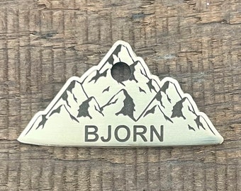 May include: A metal mountain-shaped tag with the name "BJORN" engraved at the bottom. The tag features a mountain range design with a hole at the top for attachment. The tag is silver in color and sits on a wooden surface.