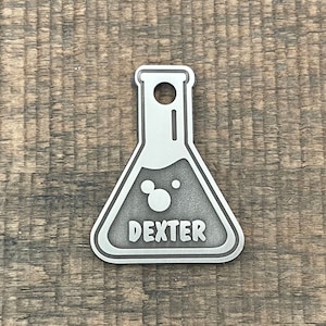 May include: Silver metal dog tag shaped like a beaker with the name Dexter engraved on it.