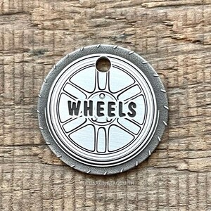 Op de afbeelding: Een zilveren metalen sleutelhanger met een wielontwerp en het woord "WHEELS" erop gegraveerd.