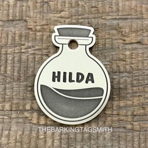 Può includere: Un'etichetta per cani in metallo argentato a forma di bottiglia di pozione con contorno nero e il nome "HILDA" inciso sul davanti. L'etichetta è su una superficie di legno.
