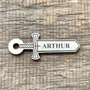 May include: A silver metal sword keychain with the name "ARTHUR" engraved on the blade. The keychain is made by "THEBARKINGTAGSMITH".