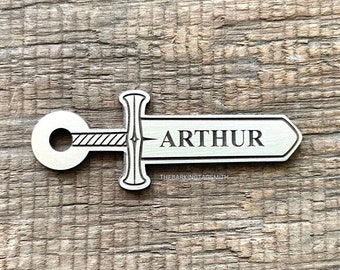 May include: A silver metal sword keychain with the name "ARTHUR" engraved on the blade. The keychain is made by "THEBARKINGTAGSMITH".