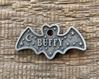 May include: A silver-toned metal pet tag shaped like a bat with a textured surface. The tag features a crescent moon, multiple stars, and the name BUFFY engraved in the center. A circular hole is at the top for attachment. THEBARKINGTAGSMITH is printed below.