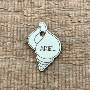Op de afbeelding: Zilveren hondenpenning in de vorm van een schelp met de naam "ARIEL" erop gegraveerd.