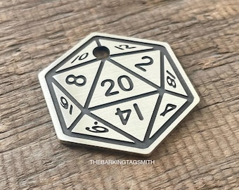 May include: A silver-colored, twenty-sided die pendant with black numbering. The numbers are arranged around the edges of the die, with the number 20 prominently displayed in the center. The pendant has a small hole at the top for a chain. The text "THEBARKINGTAGSMITH" is at the bottom.