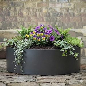 Puede incluir: Una jardinera grande, redonda, de color gris oscuro, llena de una variedad de plantas y flores. Las flores son principalmente pensamientos morados y amarillos, con follaje verde y enredaderas colgantes. La jardinera está sobre una superficie de ladrillo.