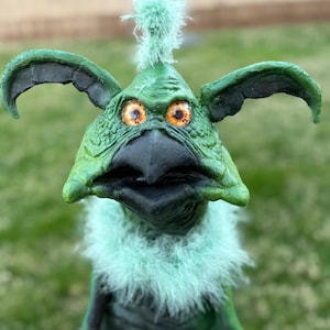 May include: A green creature sculpture with large, expressive eyes and a beak-like mouth. The sculpture has large ears and a feathery collar. The creature's skin is textured, and it has a tuft of green fluff on its head.