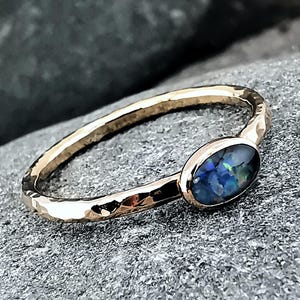 May include: A gold ring with a hammered band and an oval opal. The opal has blue, green, and black hues. The ring is set on a grey, textured surface. The ring's design is simple and elegant.
