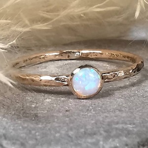 May include: A gold ring with a hammered band and a round, iridescent opal stone. The ring is set against a soft, neutral background. The ring is a beautiful piece of jewellery. The opal stone is a lovely feature.
