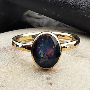 May include: A gold ring with a hammered band and a large, oval black opal gemstone. The opal displays a vibrant play of color with flashes of blue, green, and red. The ring is set on a light-colored stone surface.