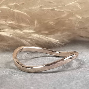 Könnte beinhalten: Ein gehämmerter Goldring mit einem wellenförmigen, asymmetrischen Design. Der Ring ist aus glattem, poliertem Metall gefertigt und hat einen dezenten Glanz.