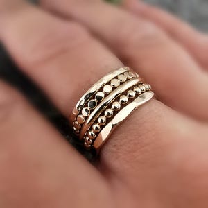 May include: Four gold rings of varying designs stacked on a finger. The rings feature hammered, beaded, and plain bands. The rings are a warm gold colour, reflecting light. The rings are a stylish accessory.