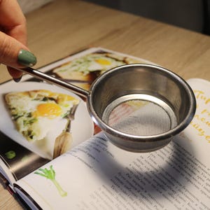 Puede incluir: Un colador de malla de plata con un mango largo. El colador está sobre un libro de cocina con una receta de una tarta salada con un huevo en el centro.