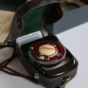 May include: A vintage black and brown leather case with a silver and red light meter inside. The light meter has a white face with the text "Leningrad 2" in red.